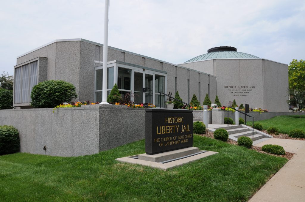 A gray building with a green front lawn. The building and the platform beneath it are made of granite. The building is made of two parts: A long section ending with a large, circular section. Several flowers and bushes sit in front of the building. A sign on the building reads “Historic Liberty Jail The Church of Jesus Christ of Latter-day Saints.