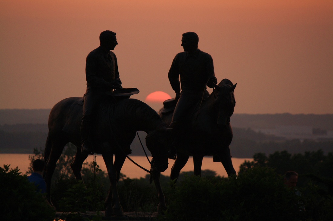 A silhouette of a statue of Joseph and Hyrum on horses. Visible in the background are trees, a river, trees on the opposite shore, and an orange sunset. The whole photo, including the river, is tinted in orange because of the sun.