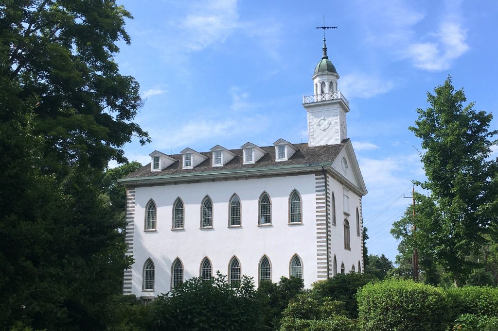 The Kirtland Temple, a white building with two stories and a garret and bell tower, is seen from across a lawn, bushes and trees.