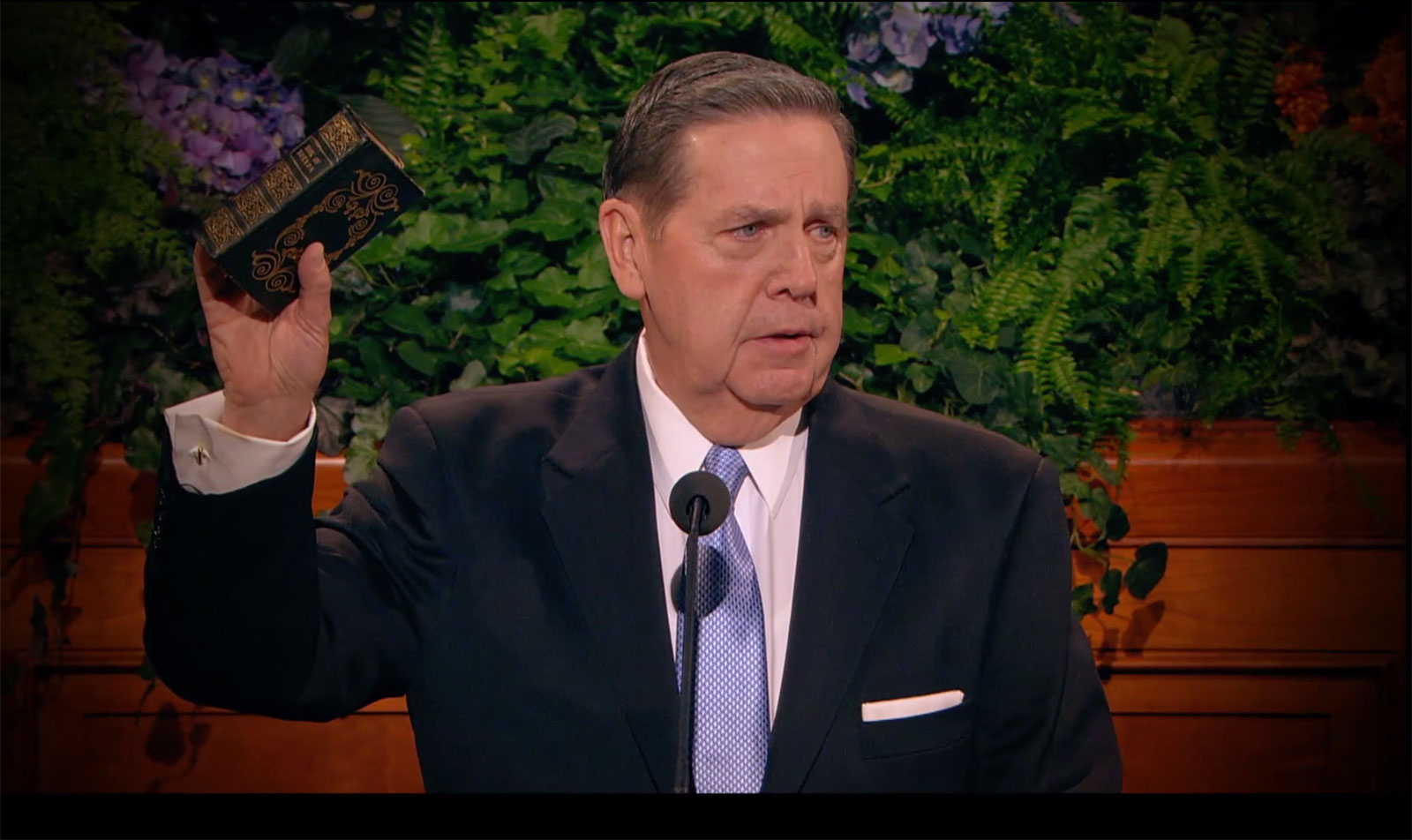 Jeffrey R. Holland, in a black suit, white shirt and light blue tie, stands at the pulpit in the conference center and holds up a copy of The Book of Mormon once owned by Hyrum Smith.