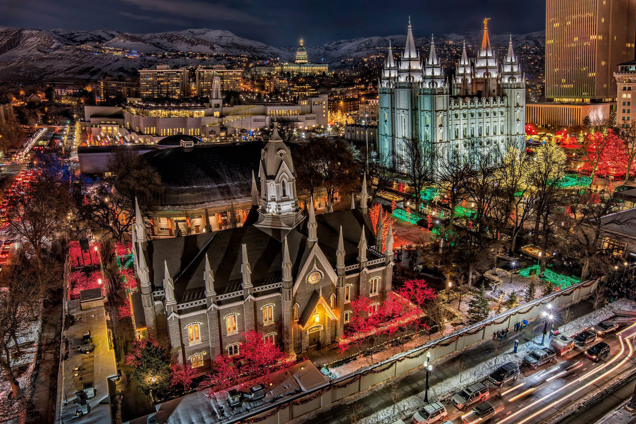 An image of Temple Square at Christmas. In the foreground is the chapel on temple square, and in the background is the temple. All around in the trees and bushes are bright Christmas lights. Other Salt Lake City building are visible in the background behind the temple.